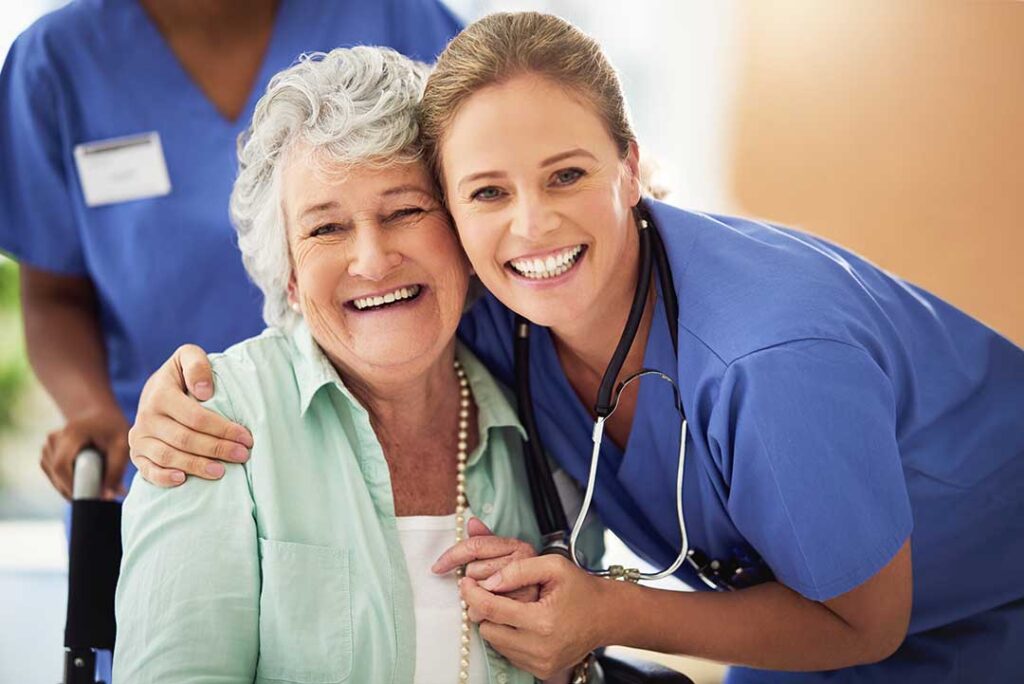 Two older adults chatting with a caregiver in a sunny common room filled with personal items and comfortable seating.